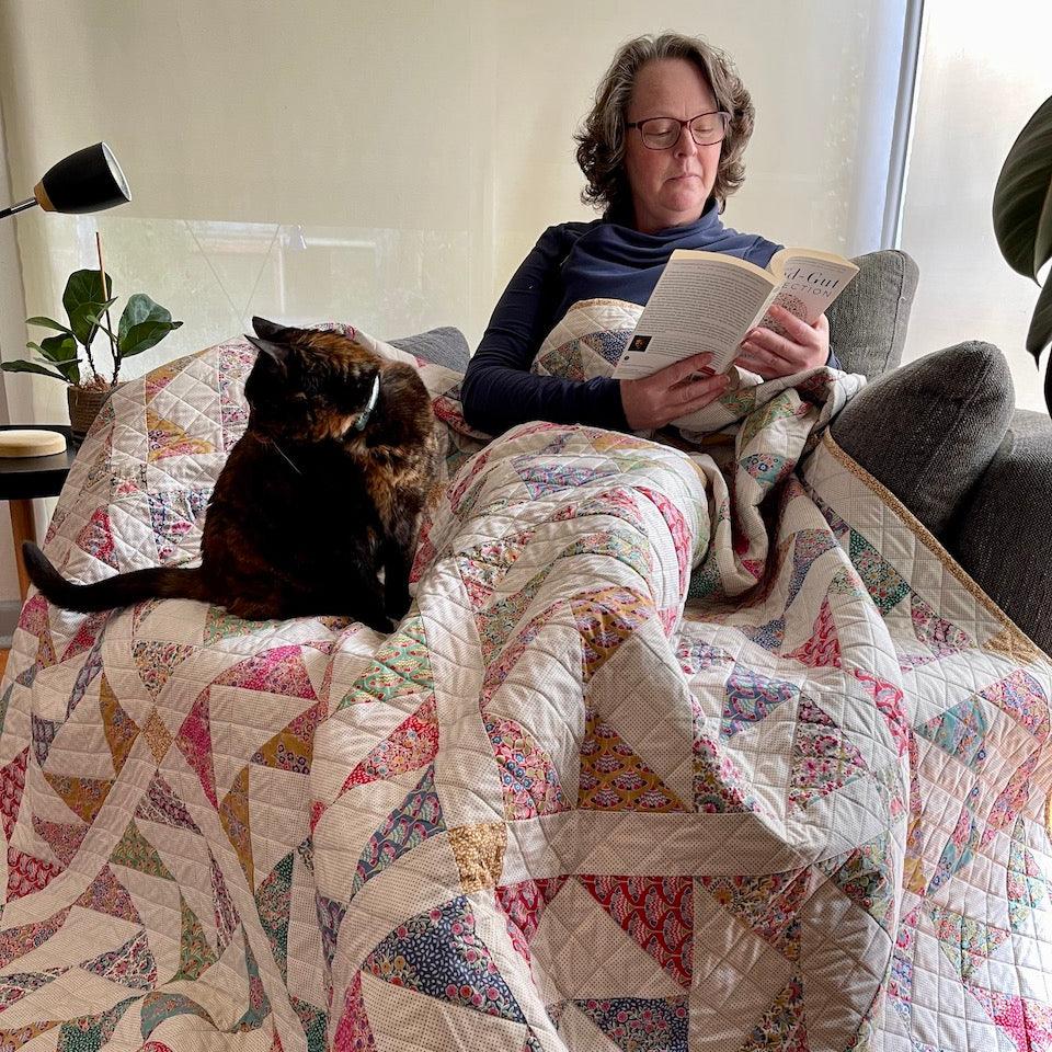 Photo of Karen, the quilt designer, sitting on a couch and reading a book with the quilt draped over her lap. The cat Lucy sits beside her.