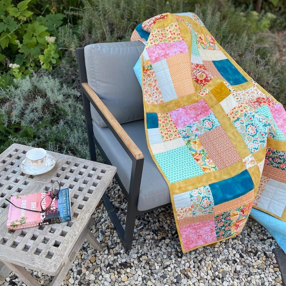 Fat Quarter quilt draped over a chair in the garden. There is a table with a book, reading glasses and a cup of tea.