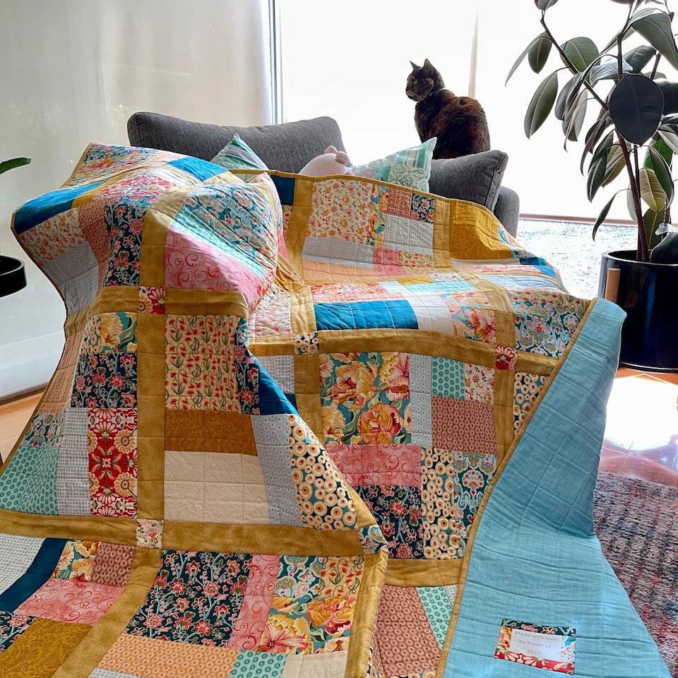 Fat Quarter Quilt draped over a couch with a corner of the quilt turned over to show the quilt backing. Lucy the cat sits in the background on the arm of the couch.
