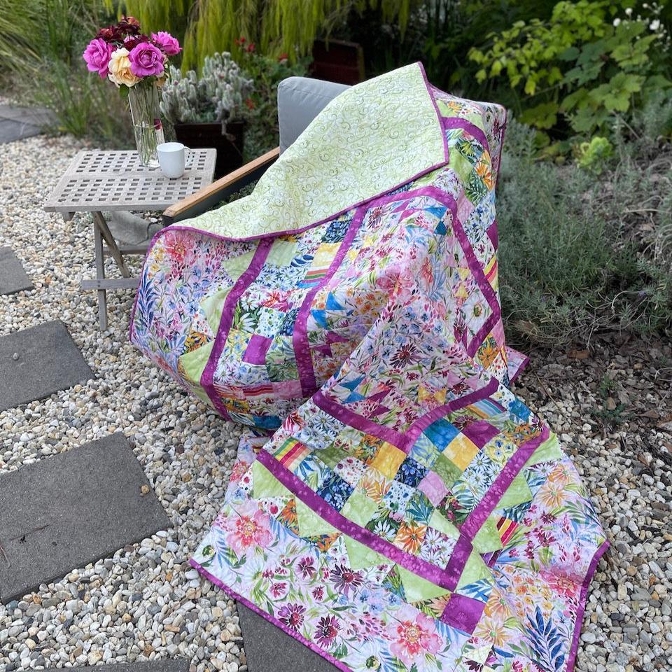 My Mother's Garden Quilt draped over a chair in the garden. Stepping stones in the foreground and garden in the background. Beside the chair is a table with a coffee cup and a vase of pink and yellow roses.