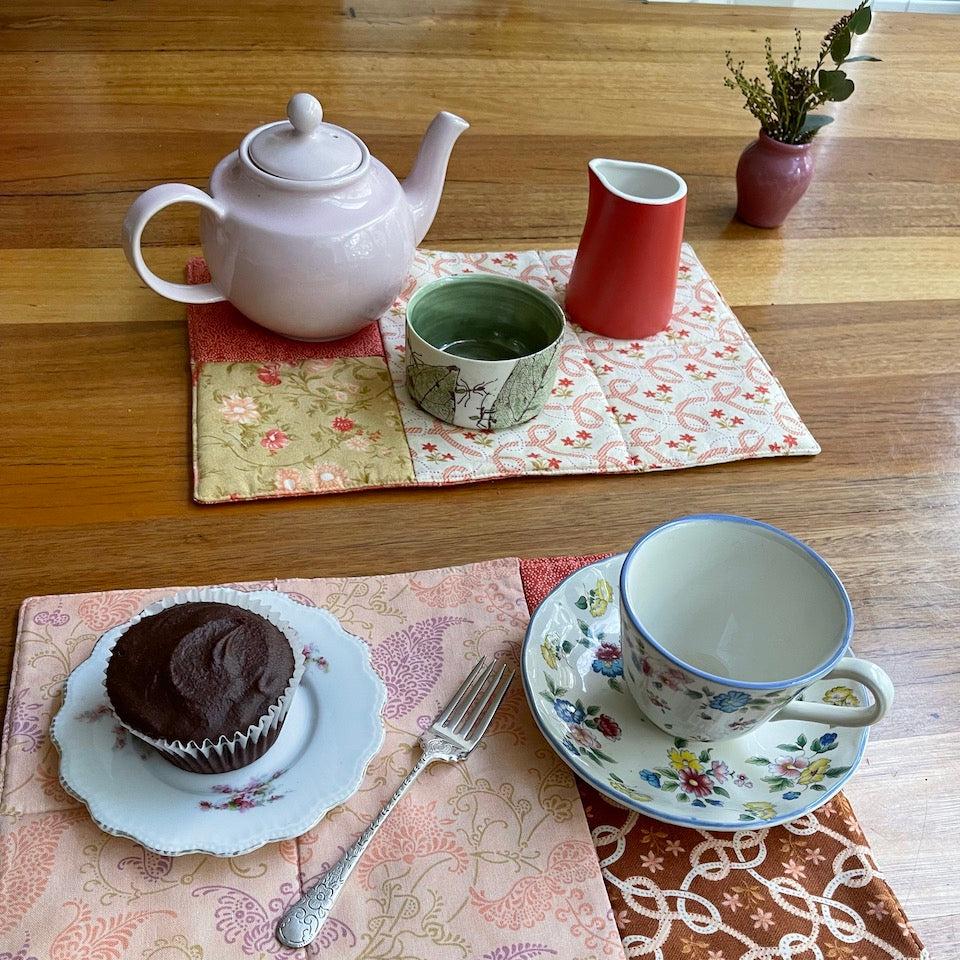 Placemats with a cupcake on a plate with a fork, tea cup and saucer. in the background are flowers, a tea pot and milk jug.