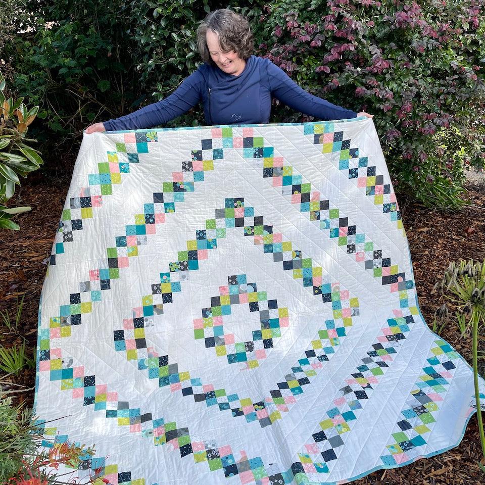 Rough Diamond Quilt being held up by Karen, Quilt Designer. The bottom of the quilt is on the ground as she looks down at the quilt. There is garden in the background. The coloured fabrics look like little squares pieced together to form diamond shapes radiating outwards. Each diamond is bigger than that one before it.