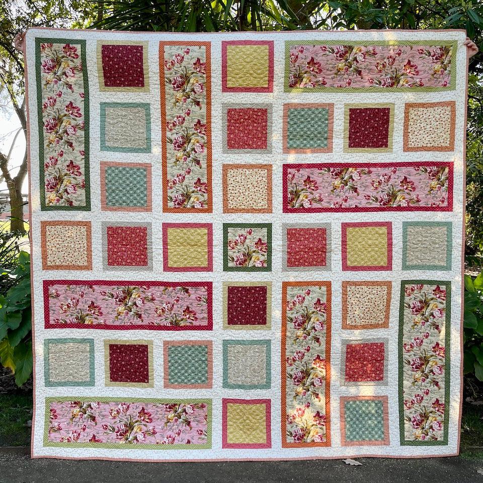 The Tulip Quilt held up in front of a garden. The quilt is made of Tulip themed fabrics. The Tulip fabrics are surrounded by small colourful borders. Some of the quilt blocks are square and others large rectangles.
