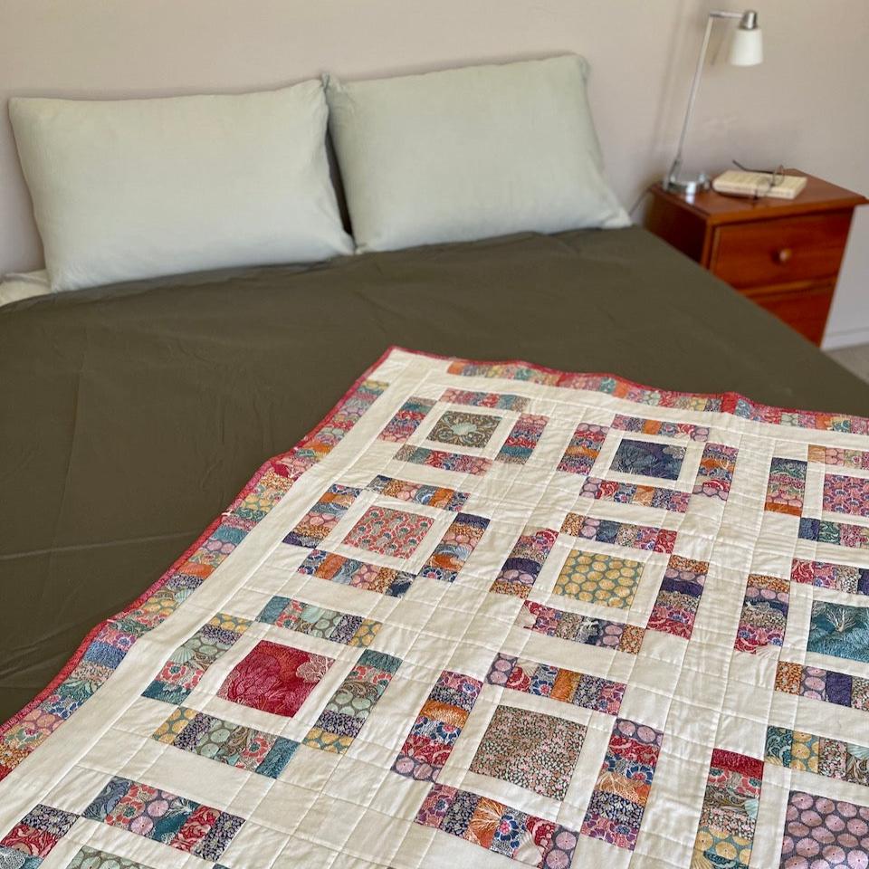 Anemone Quilt on an angle across a bed.  The bed has soft sage green pillows and a grey green cover with the quilt over the top.  There is also a bedside table.