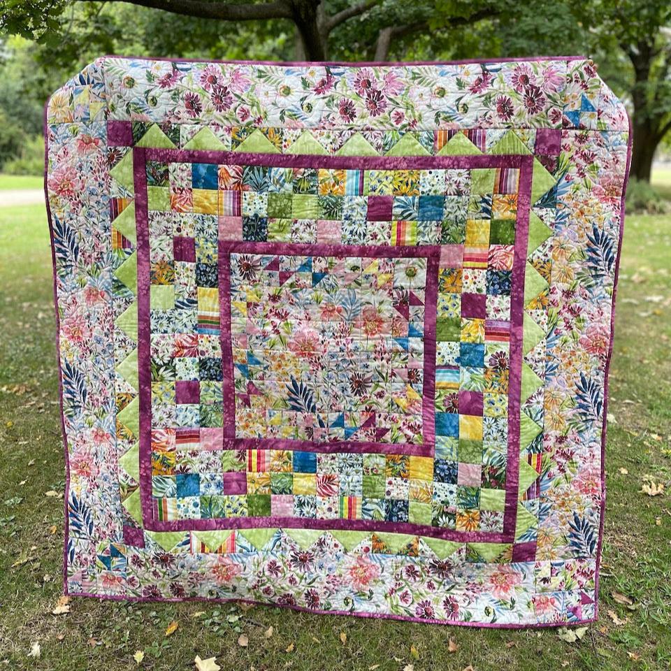 My Mother's Garden Quilt being held up in front of a tree.  The quilt is in floral and solid fabrics.  It has a centre square of floral fabric surrounded by a number of different pieced borders in coordinating colours and prints.