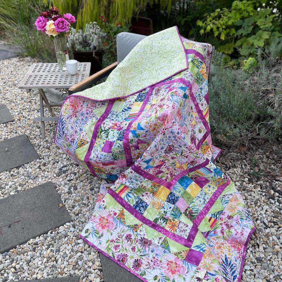 My Mother's Garden Quilt draped over a chair in the garden.  Stepping stones in the foreground and garden in the background.  Beside the chair is a table with a coffee cup and a vase of pink and yellow roses.