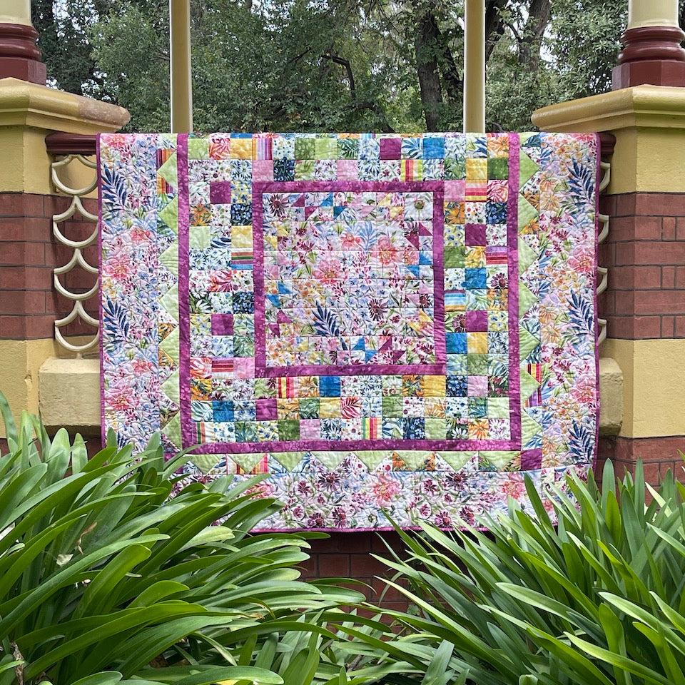 My Mother's Garden Quilt hanging over the handrail of a bandstand with garden in front and tree tops in the background.