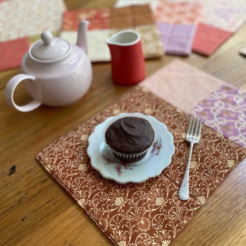 A placemat in brown, purple, pink and cream with a cake a delicate plate, cupcake and fork.  there are other placemat with a tea pot and milk jug in the background. 