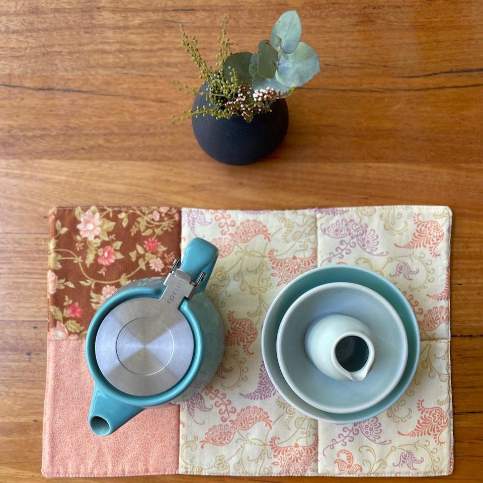 A placemat on a wooden table with a tea pot bowls and milk jug on top taken from above. There is a small vase of flowers at the top of the picture and a blue rug in the bottom fading into the background.