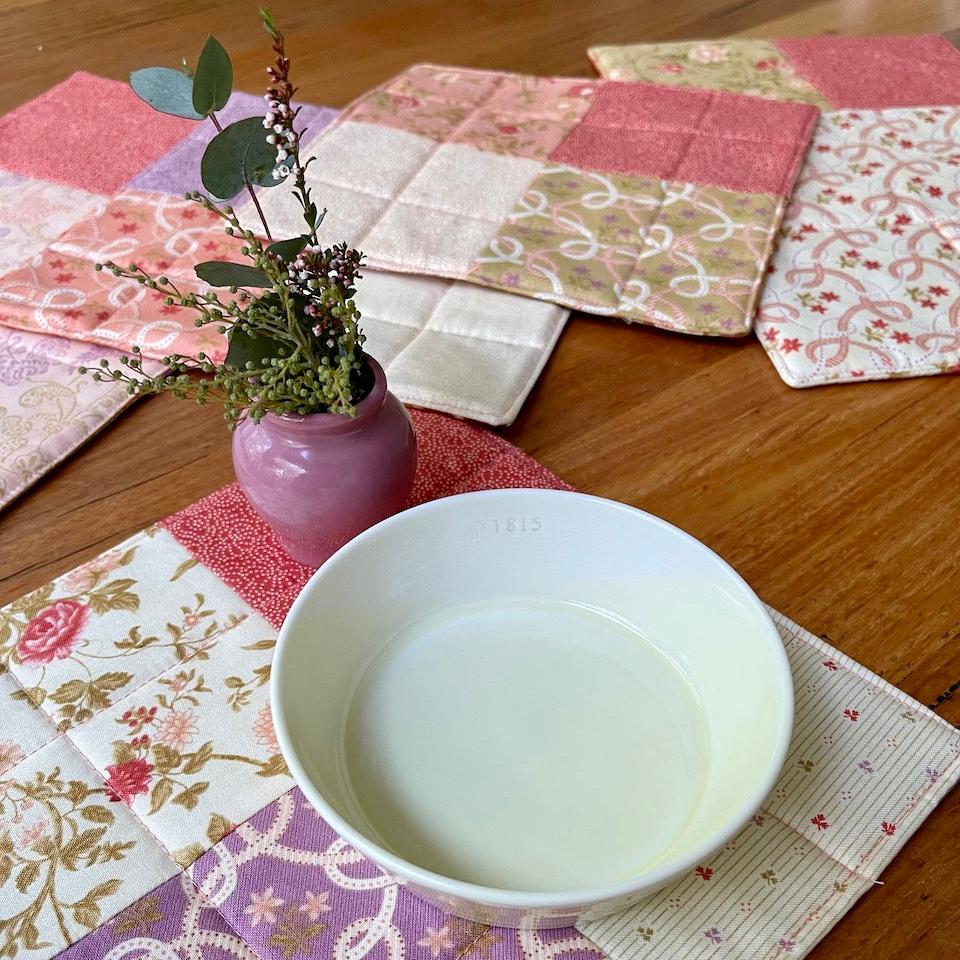 Mug rugs and placemats laid on a wooden table with a small ceramic bowl and a small vase of flowers.  The placemats are in salmon pinks, purples, green and cream.