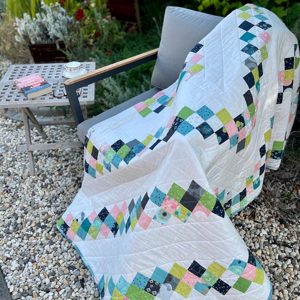 Rough Diamond Quilt draped over a chair in the garden.  There is a table beside the chair with a book, reading glasses and a tea cup.