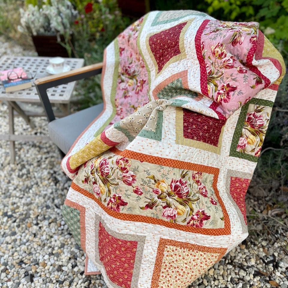 Tulip quilt draped over a chair in the garden.  There is a table with a book, reading glasses and a cup of tea beside it.